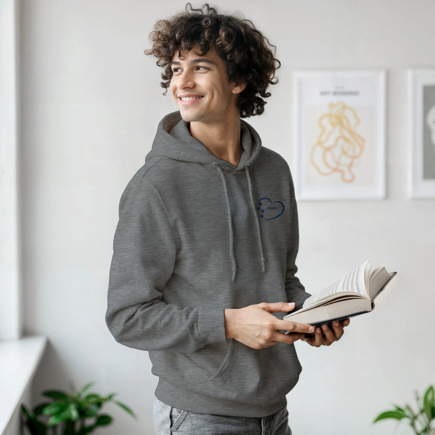 Custom Embroidery, Heart Personalized with Your Pet's Name on a Pullover Grey Hoodie Sweatshirt hearts inside blue thread young man standing in a room holding a book and smiling.
