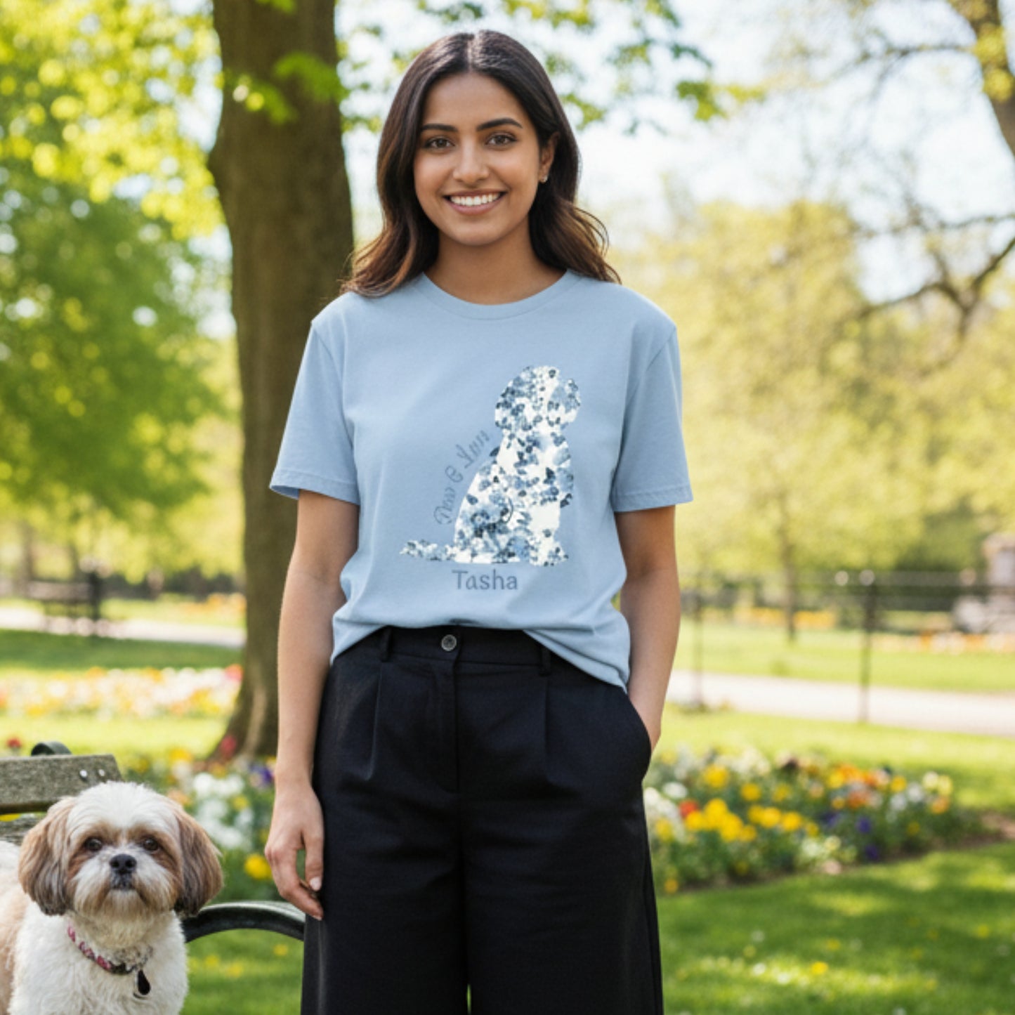 Woman wearing a light blue personalised t-shirt with a toile de jouy coquette floral Shih Tzu shaped graphic and text 'Paws & Love' in a park with a dog on a bench.