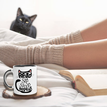 Black cat aesthetic ful mug with black inside and a cat image with text reading 'read, sip, sleep, repeat.' In the background a person wearing socks and sleeping next to a book. 