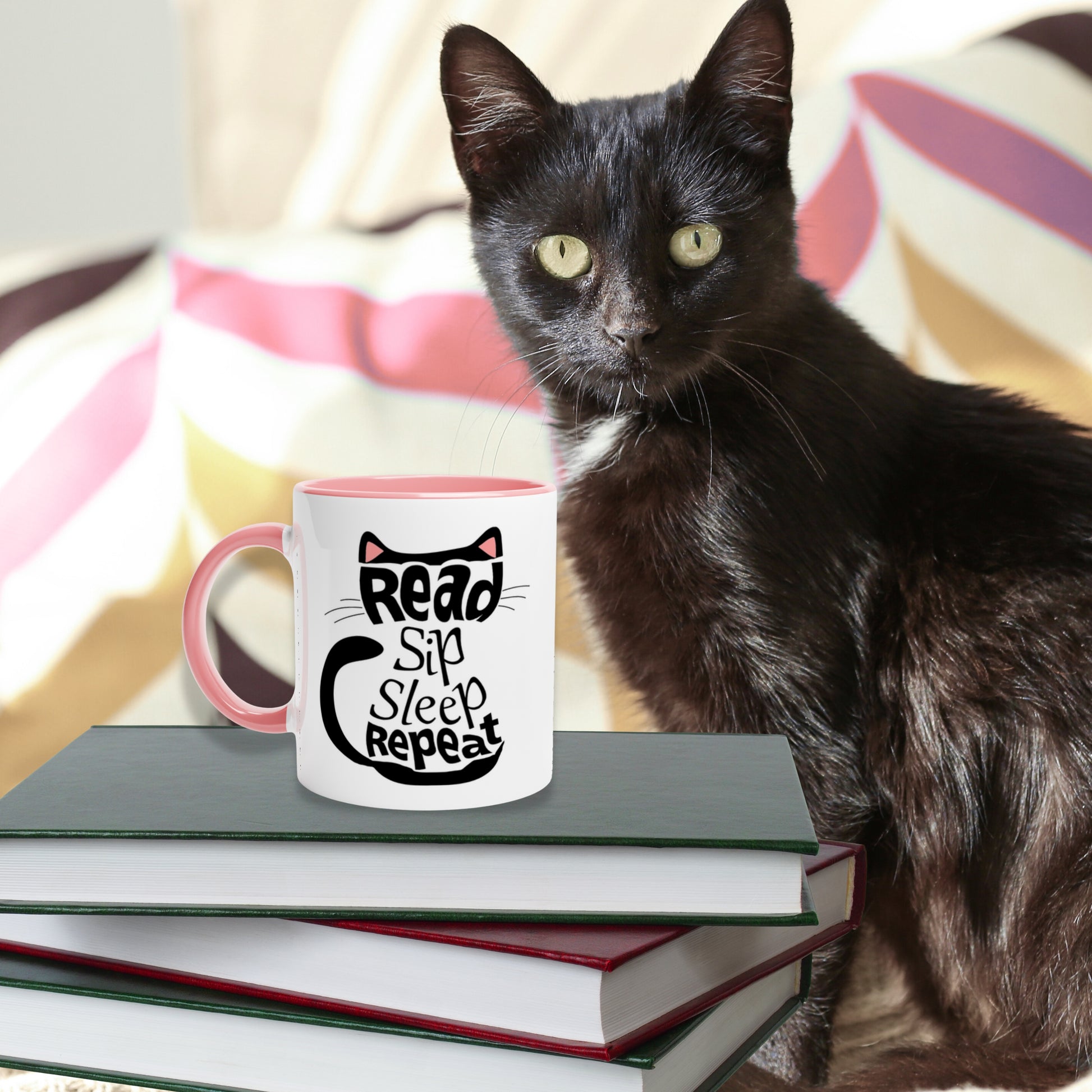Black cat sitting next to a stack of books.On top of them is a pink color inside mug with 'Read Sip Sleep Repeat' text inside of a black cat with pink ears.