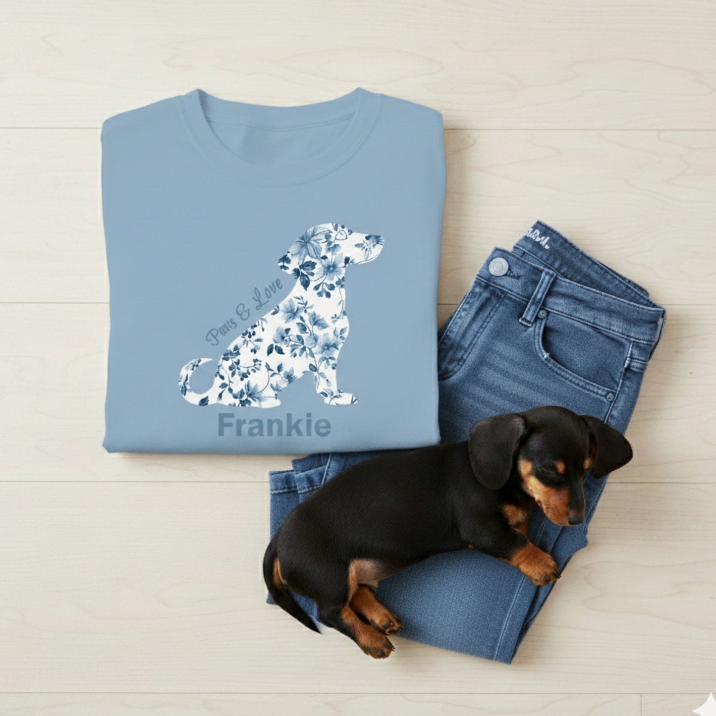 Folded light blue t-shirt with floral dachshund design and text with dog's name on the tee, jeans, and a small black and brown dachshund puppy on a light wooden surface.