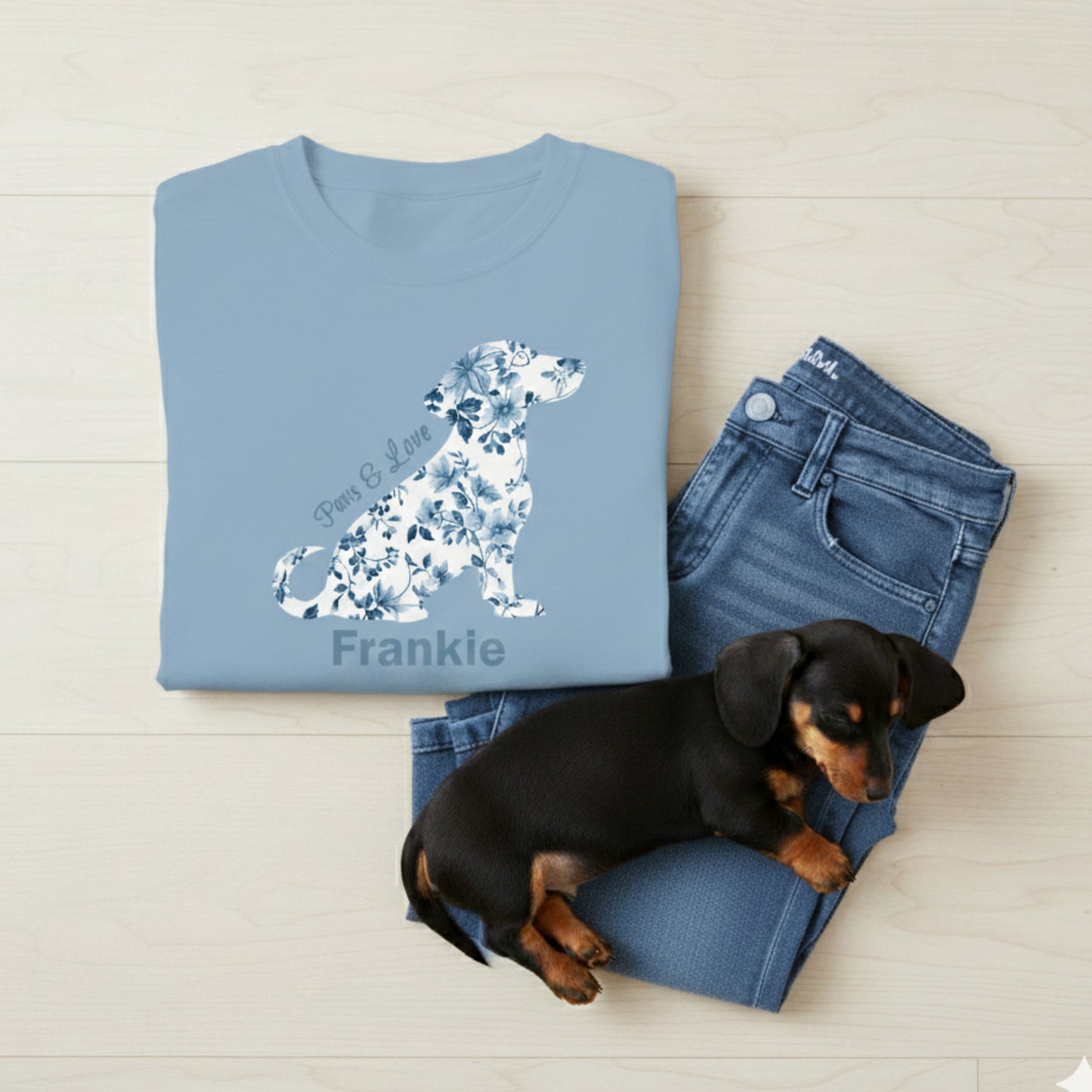 Folded light blue t-shirt with floral dachshund design and text with dog's name on the tee, jeans, and a small black and brown dachshund puppy on a light wooden surface.