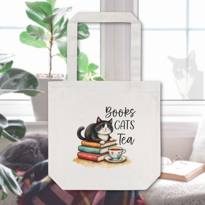 Tote bag with tuxedo cat on top of books in front of a tea cup in the background is a cozy reading space with a cat looking in the window.