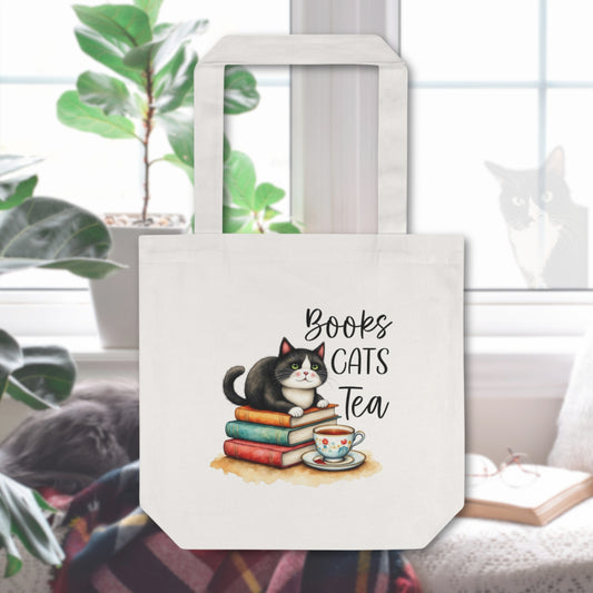 Tote bag with tuxedo cat on top of books in front of a tea cup in the background is a cozy reading space with a cat looking in the window.