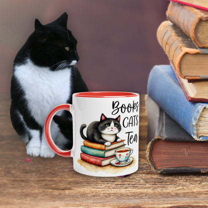 Tuxedo cat sitting next to a mug with a cat illustration and text 'Books Cats Tea' on a wooden table with books.