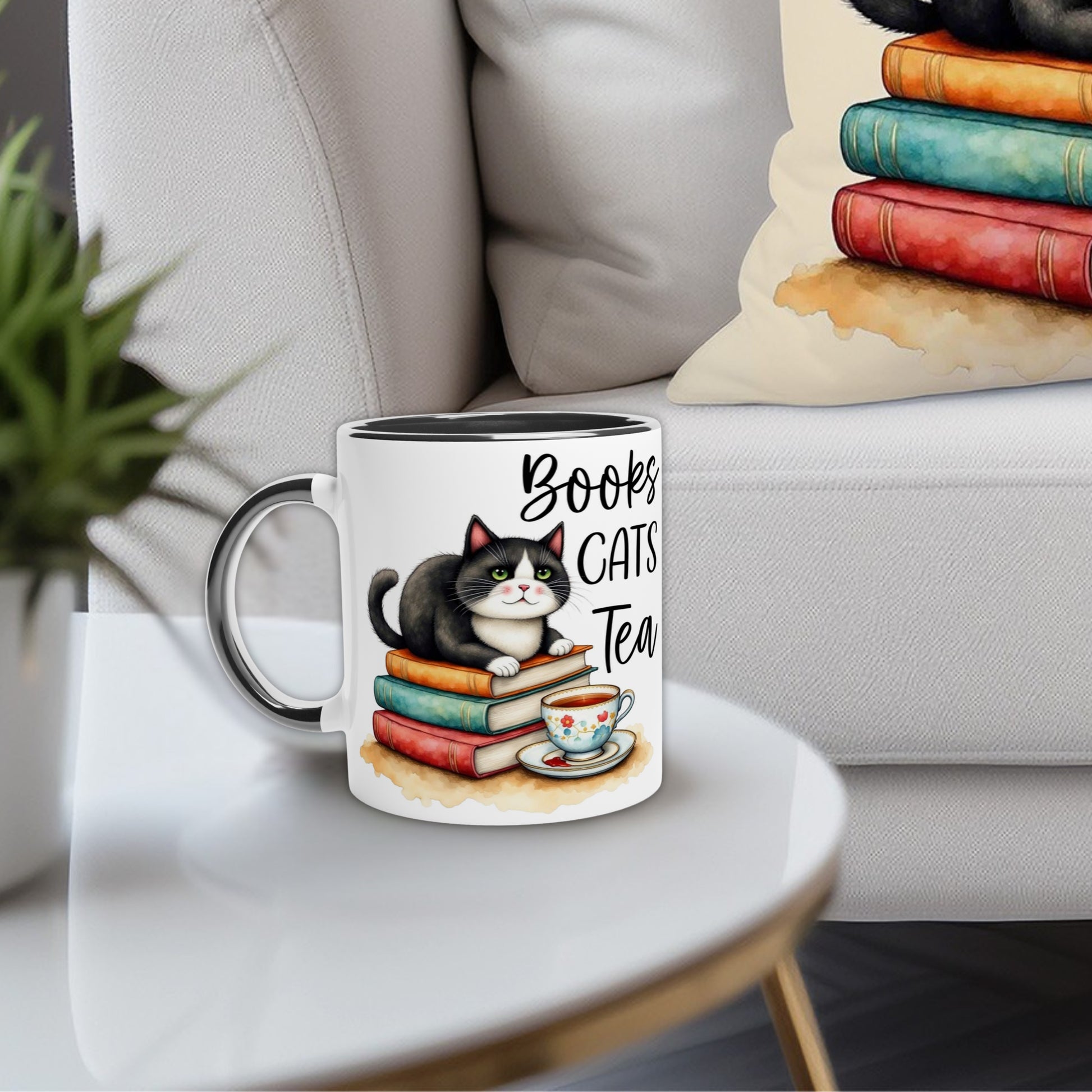 Novelty color inside mug with a cat illustration and text 'Books Cats Tea' on a table in a cozy living room setting. The pillow on the couch has the same bookish aesthetic as the tea mug
