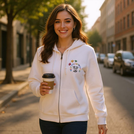 Woman in a white Zipper hoodie with a colorful embroidery design of a dog, dog paws and the words, Best Dog Mom Ever!. The woman is holding a coffee cup while walking on a city street.