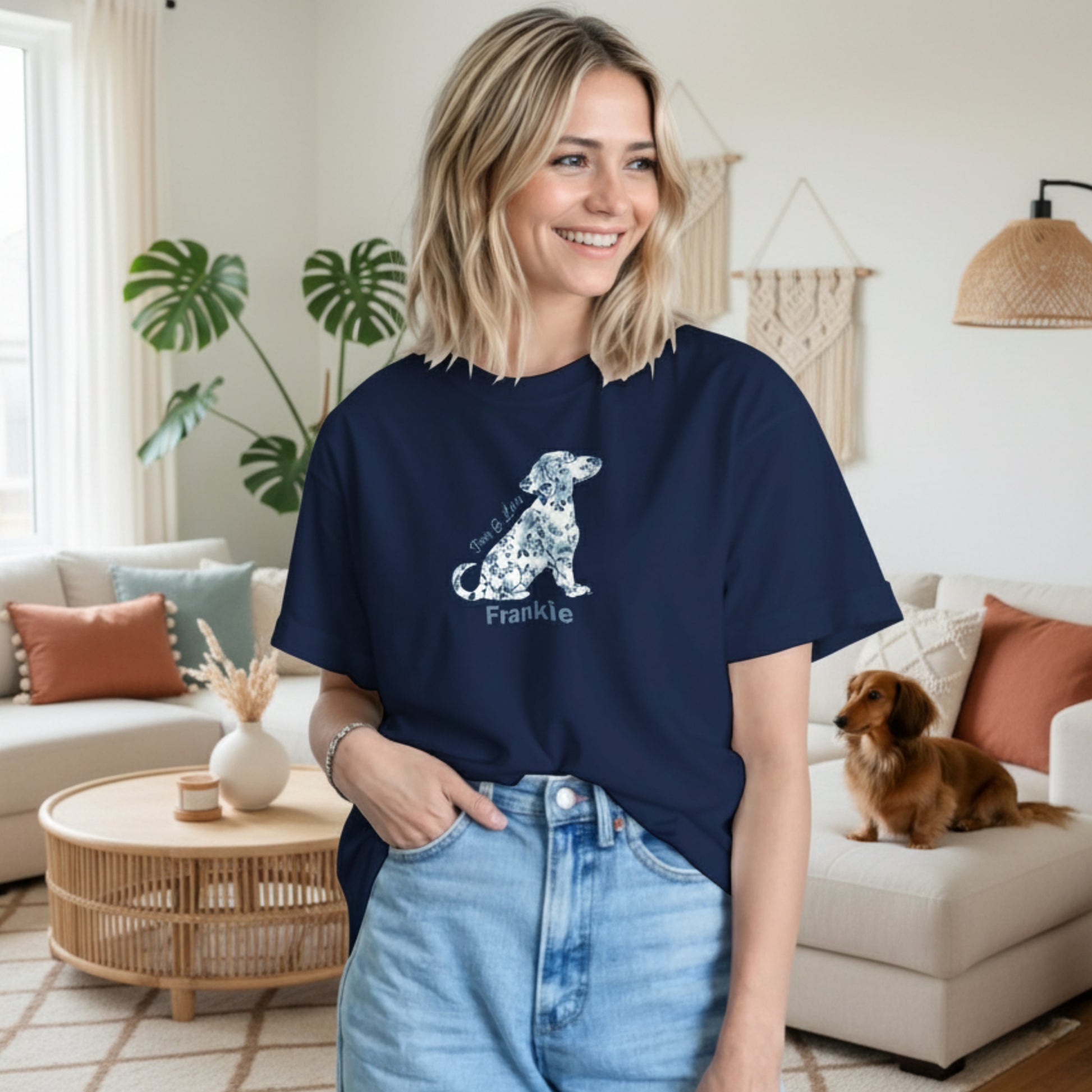 Woman wearing a navy blue t-shirt with a coquette floral pattern in the shape of a Dachshund graphic and text 'paws & Love' and the dog's name in a Boho style living room with a long haired Dachshund sitting on the couch.