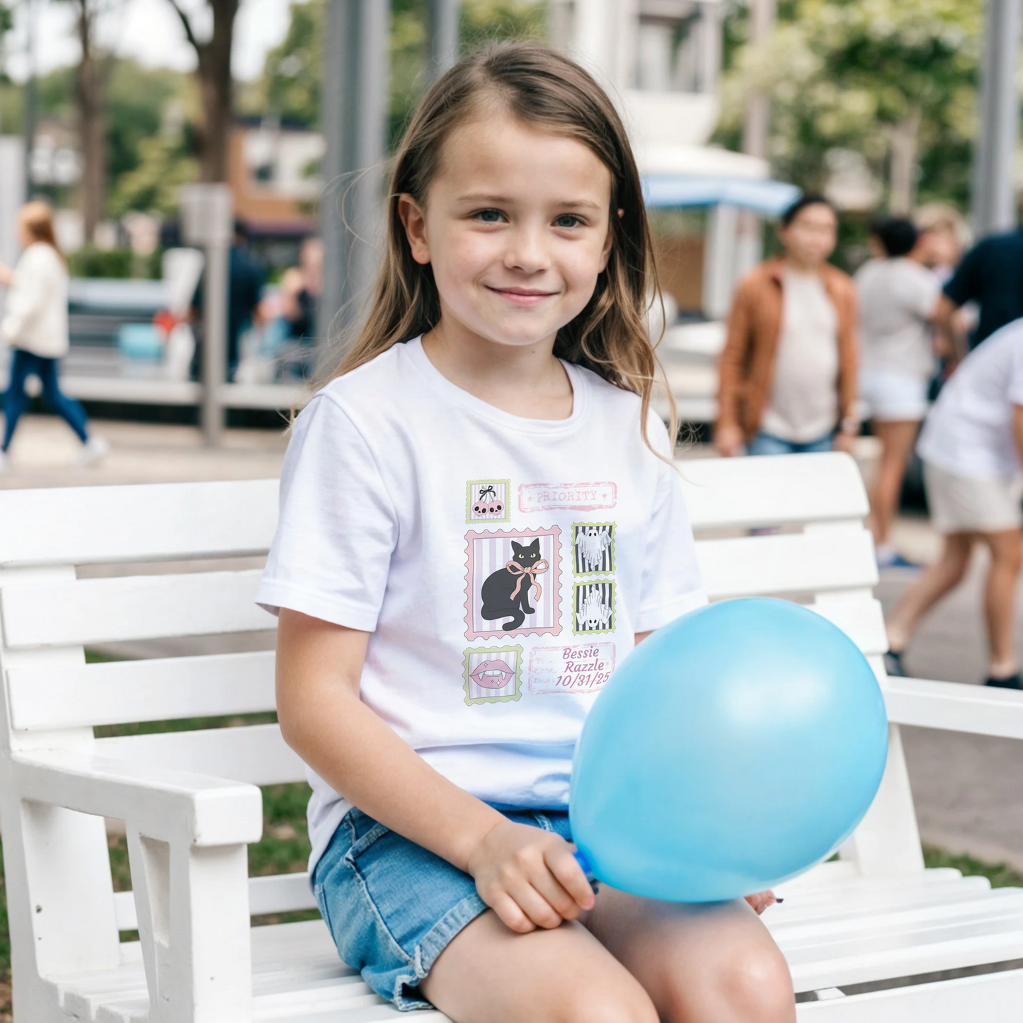 Young girl  wearing a white t-shirt with a coquette pink Halloween stamp graphic holding a blue balloon sitting on a white bench in an outdoor setting. 