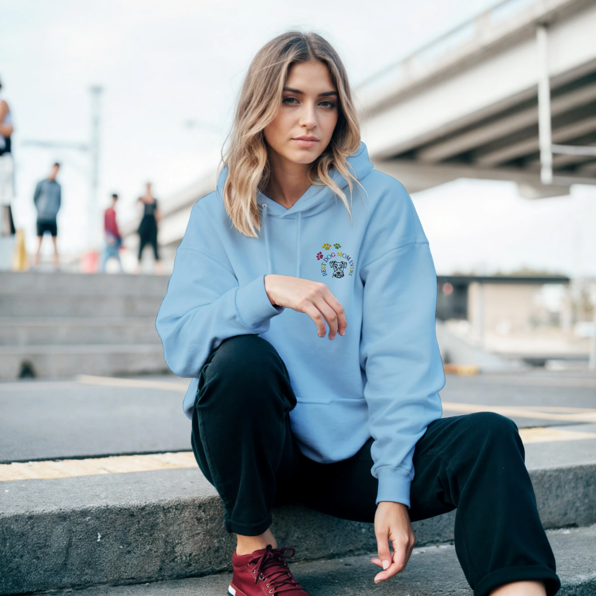 Custom Embroidered Classic Pullover Hoodie | Best Dog Mom Ever! A Woman in a light blue hoodie sitting on steps outside in a city.