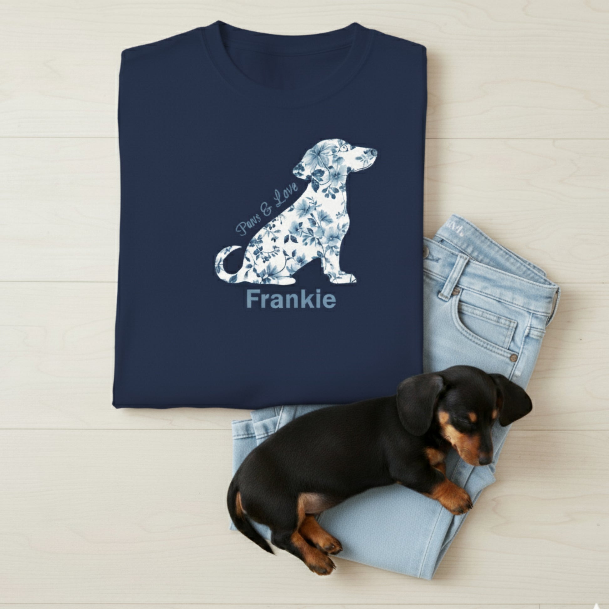 Navy cottagecore blue floral dachshund t-shirt with 'Paws & Love text and dog's name. Tee is folded lying on light wood surface with a puppy sleeping on jeans.