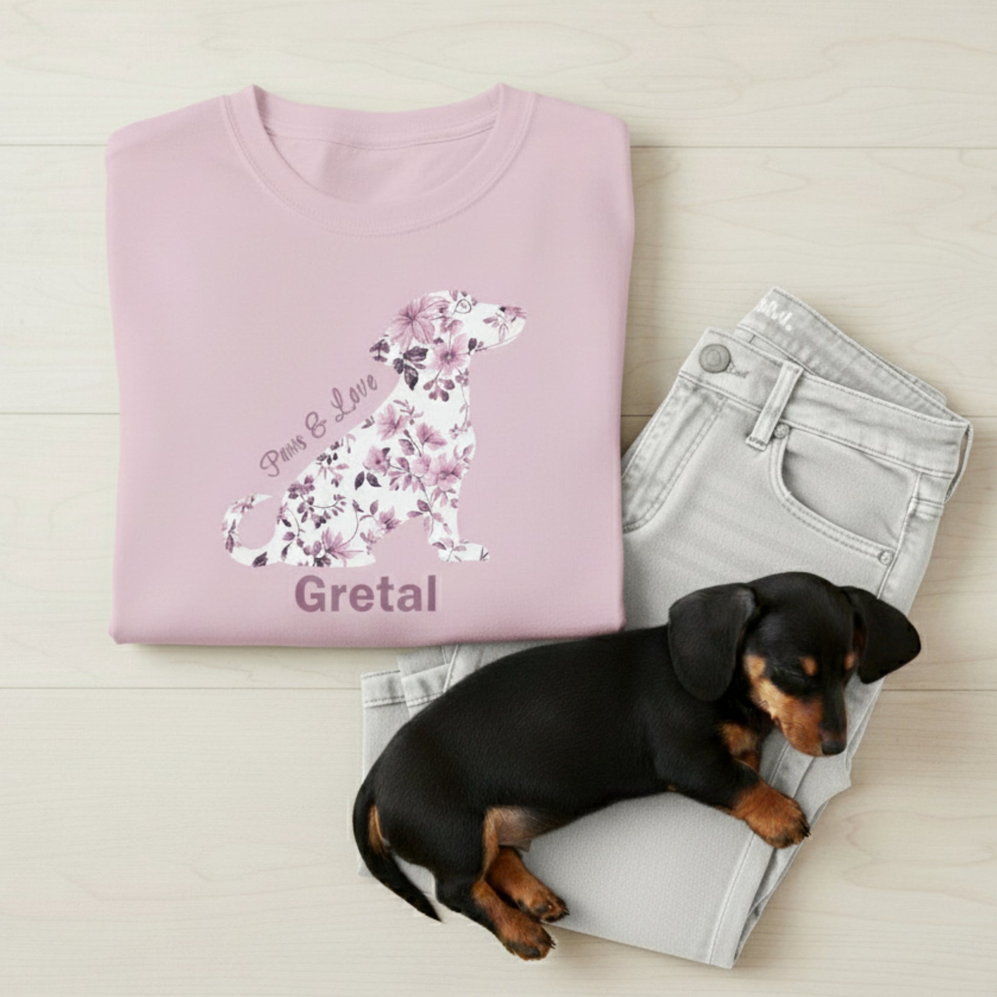 Pink t-shirt with toile de jouy floral dog design and 'Gretal' text for dog's name, lying next to a small black and brown dog on light-colored wood.