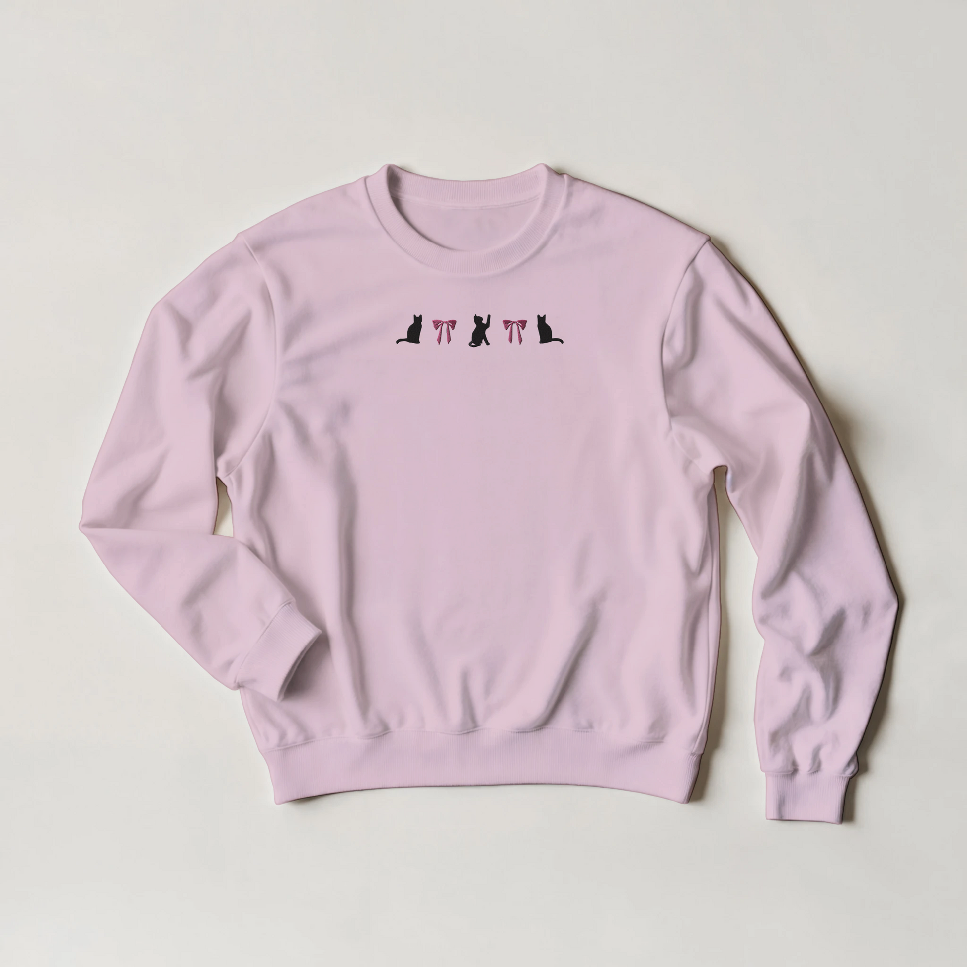 Pink sweatshirt with black cats and pink bows embroidered in a line across the front. The shirt is flat on a light gray background