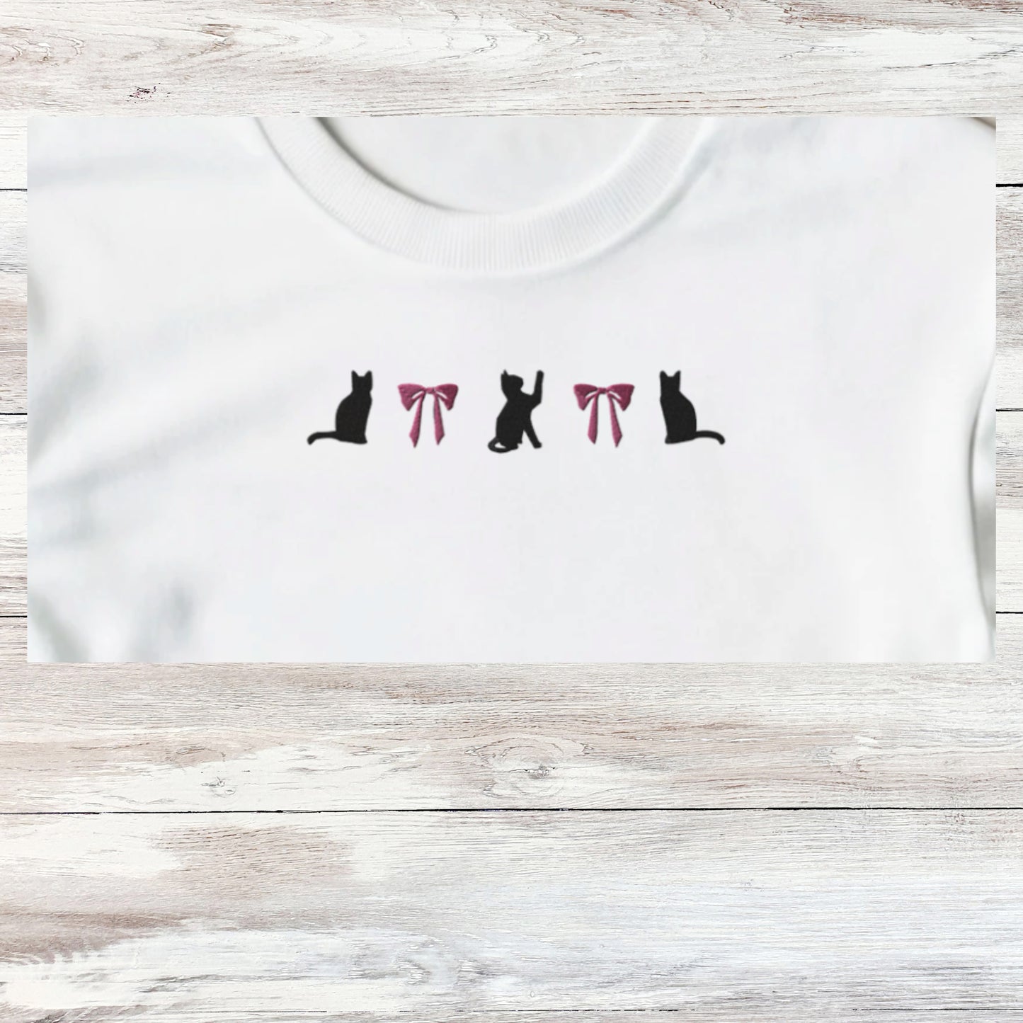 Embroidery of three playful black kittens and to coquette pink bows, design shown close up on a white sweatshirt that is folded.