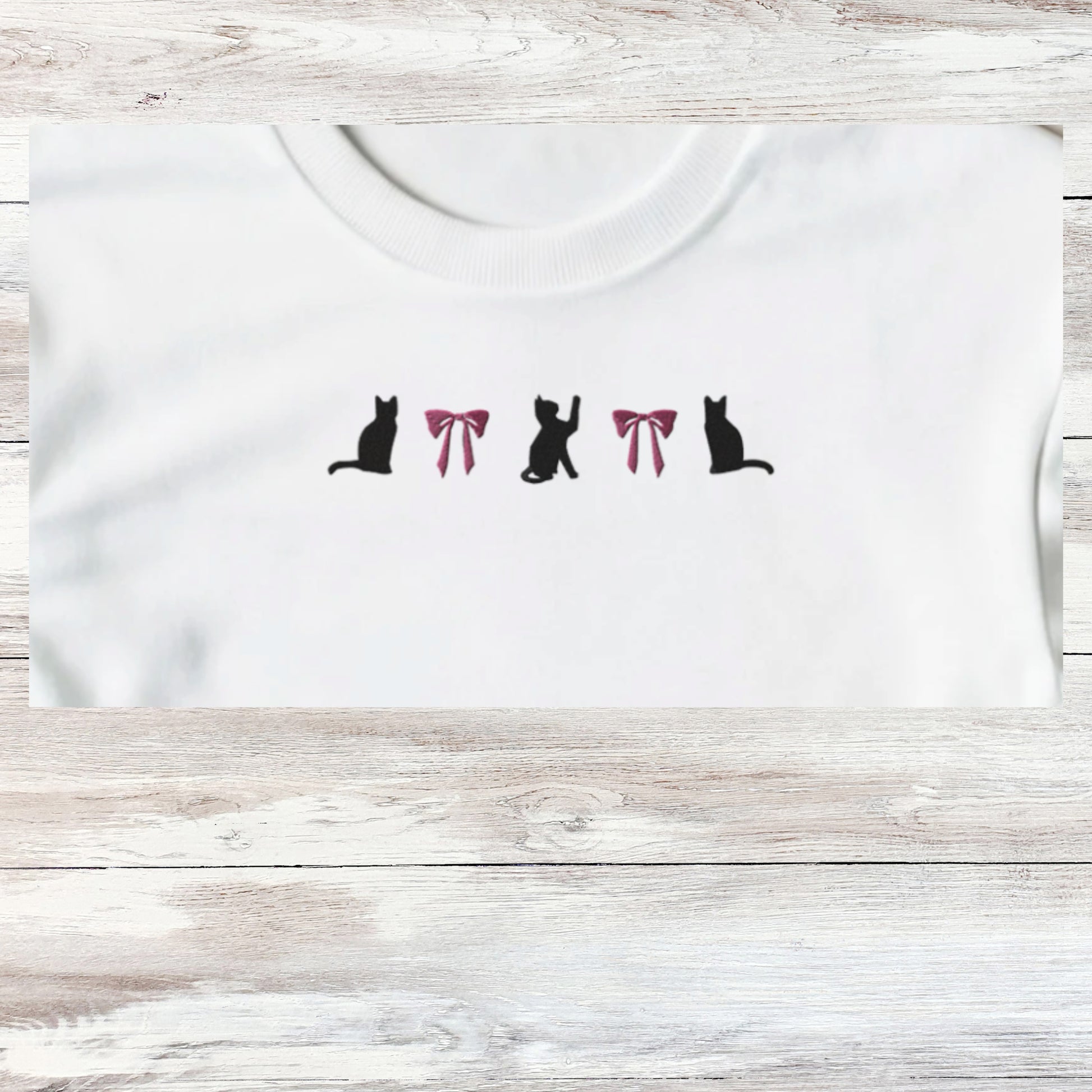 Embroidery of three playful black kittens and to coquette pink bows, design shown close up on a white sweatshirt that is folded.