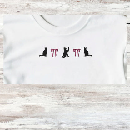 Embroidery of three playful black kittens and to coquette pink bows, design shown close up on a white sweatshirt that is folded.