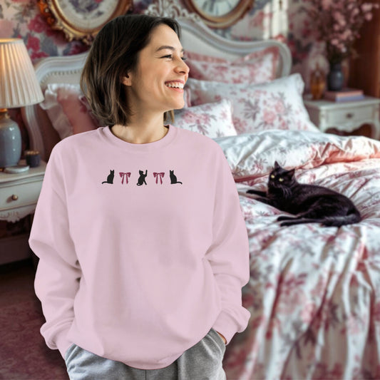Woman wearing a light pink sweatshirt with black cat and pink coquette embroidered design in a bedroom with a black cat on a bed.