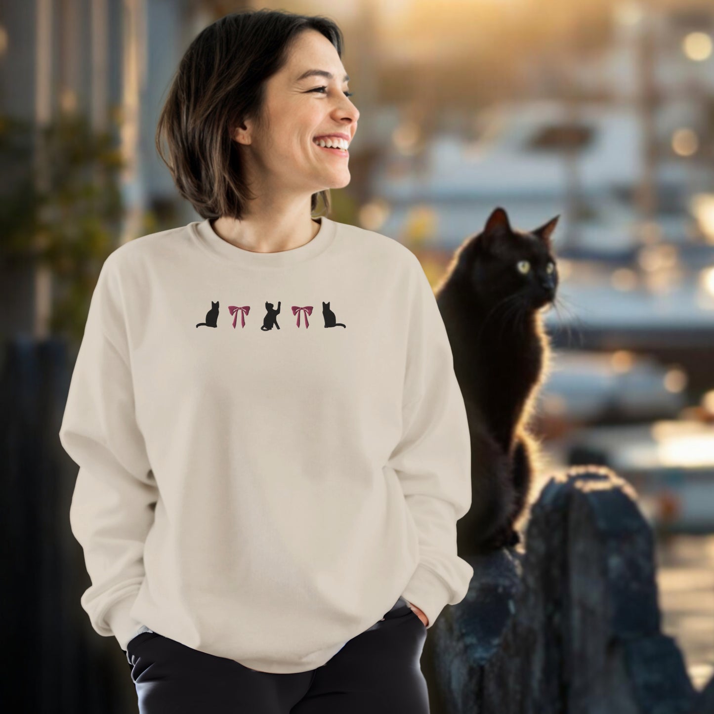 Woman wearing a sand sweatshirt with an embroidered black cat and pink coquette bows design. She is  standing next to a black cat outdoors in front of a harbor.