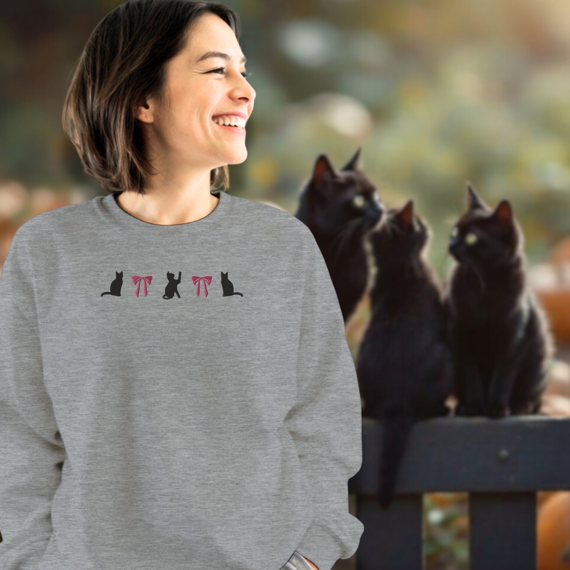 Woman wearing a sport gray sweatshirt an embroidered design of black cats and pink bows standing in front of three black cats who are sitting on a fence in front of a pumpkin patch in the late afternoon.