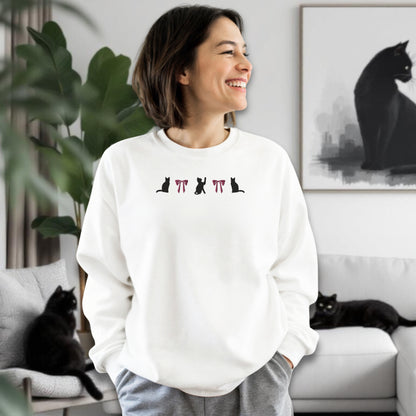 Woman wearing a white sweatshirt an embroidered black cat and pink bow design in a living room where two black cats are sitting.