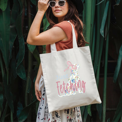 Young woman dressed up and wearing a hat carrying a tote  with a pretty Labrador wearing a pink bow on the shopping bag. Text on the bag - Just Fetching