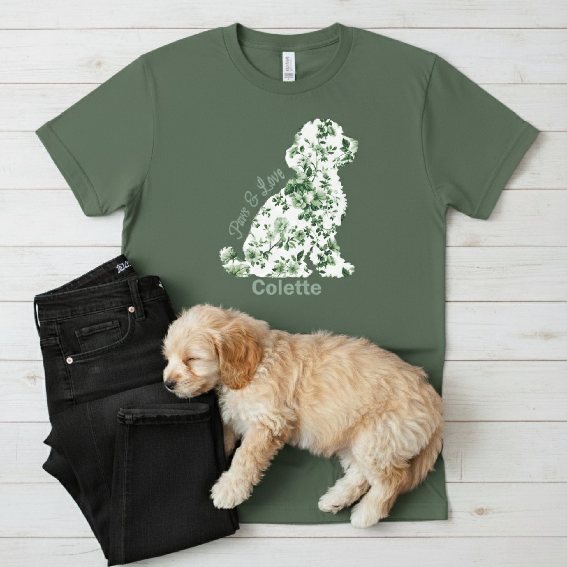 Green t-shirt with a floral cottagecore dog graphic and the dog's name next to a sleeping Cavapoo and dark jeans on a wooden surface.