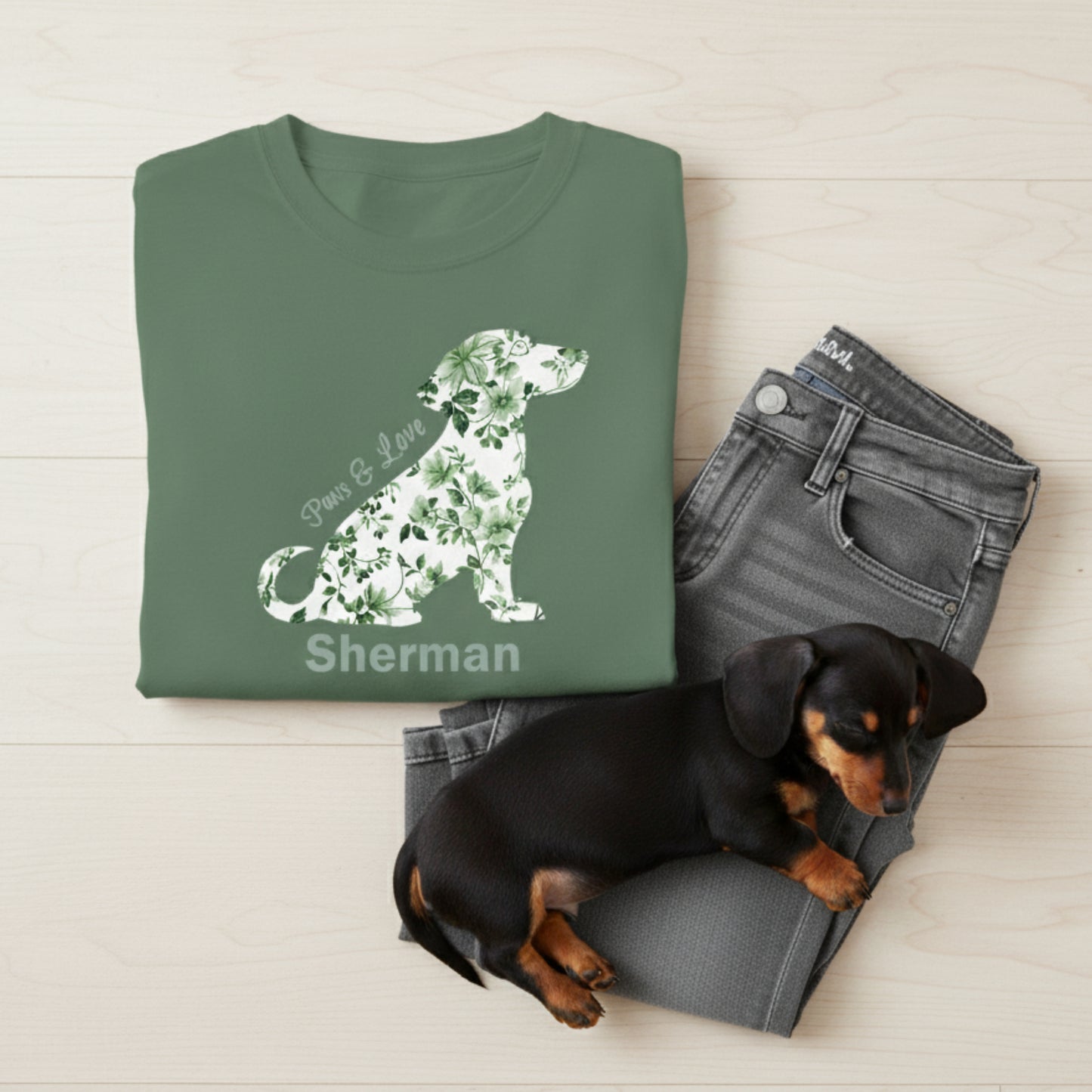Green t-shirt with Toile de Jouy floral dauchshund design and text lying on gray jeans with a small black and brown dog.