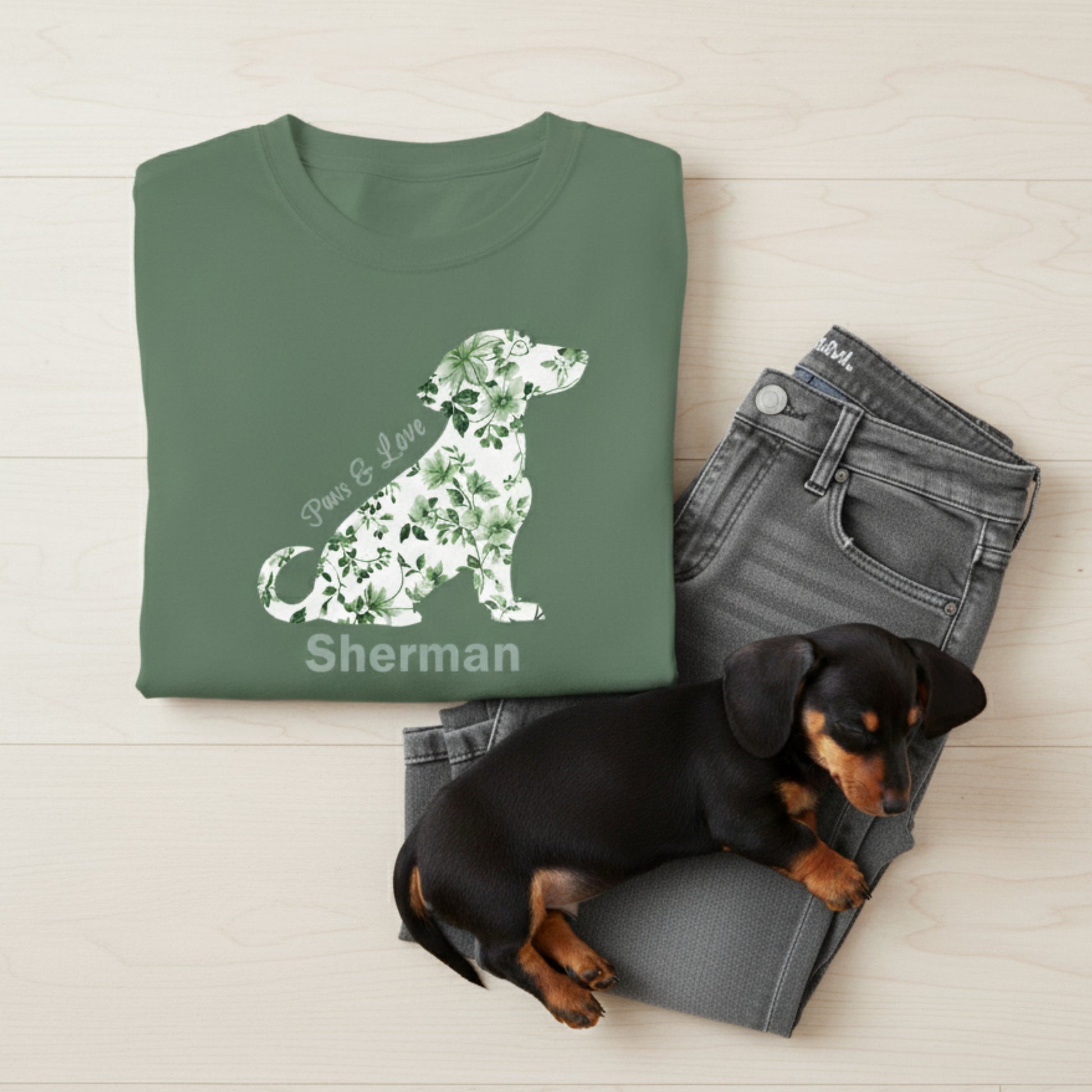 Green t-shirt with Toile de Jouy floral dauchshund design and text lying on gray jeans with a small black and brown dog.