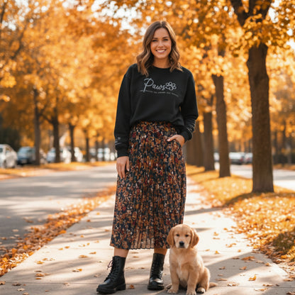 Woman in a black pullover crew neck custom embroidered sweatshirt and floral skirt standing with a puppy dog on a sidewalk lined with trees with autumn foliage.