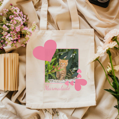 Eco Friendly cotton Tote bag with a pink heart picture frame inside the frame is a photo of a sweet little orange kitten. The bag is on top of a beige fabric background with pink flowers.