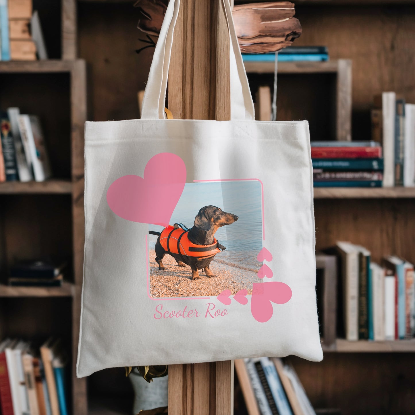 Tote book bag hanging against a bookshelf. On the tbag is a photo of a dachshund in an orange life jacket within a pretty pink hearts frame.
