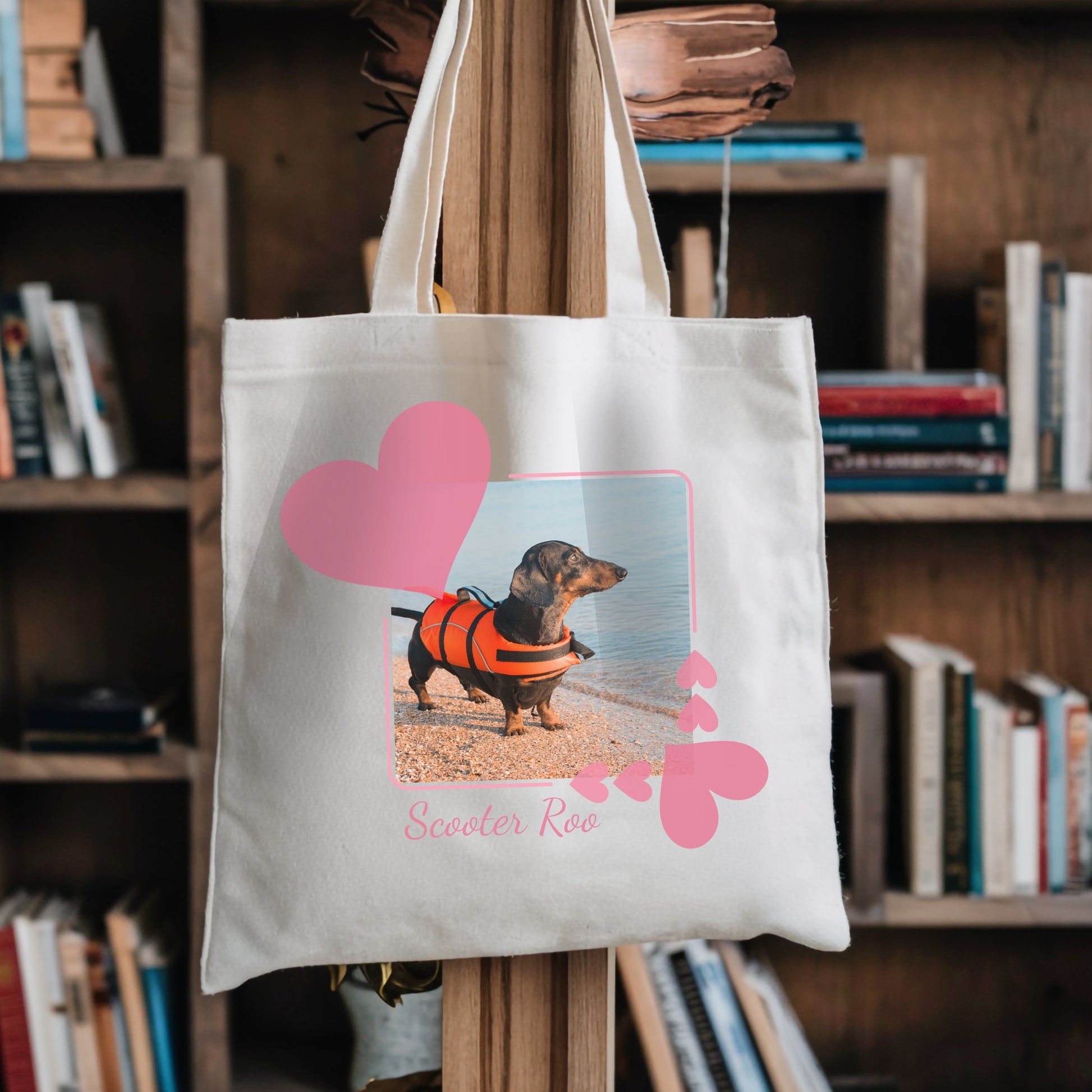 Tote book bag hanging against a bookshelf. On the tbag is a photo of a dachshund in an orange life jacket within a pretty pink hearts frame.