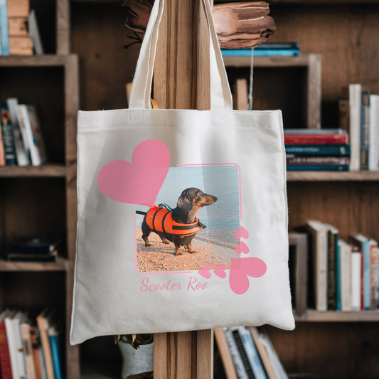 Tote book bag hanging against a bookshelf. On the tbag is a photo of a dachshund in an orange life jacket within a pretty pink hearts frame.