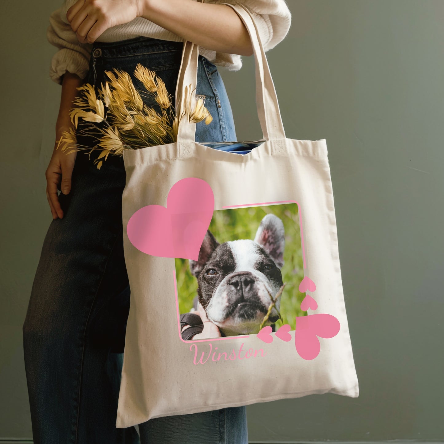 Person holding a reusable shopping tote bag with a dog photo within a frame of pink hearts on a plain background