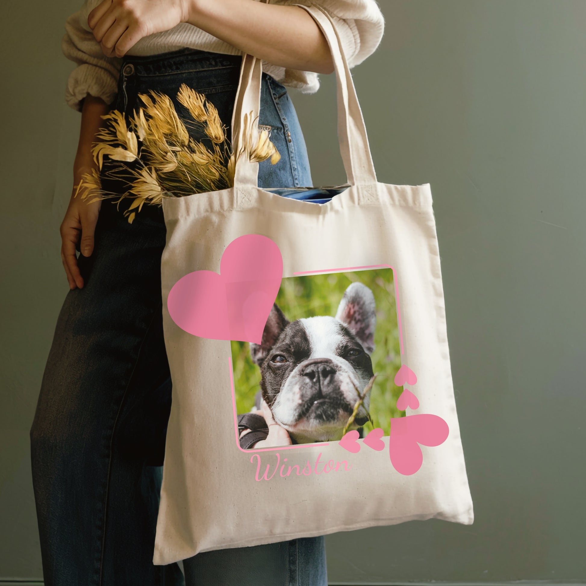 Person holding a reusable shopping tote bag with a dog photo within a frame of pink hearts on a plain background