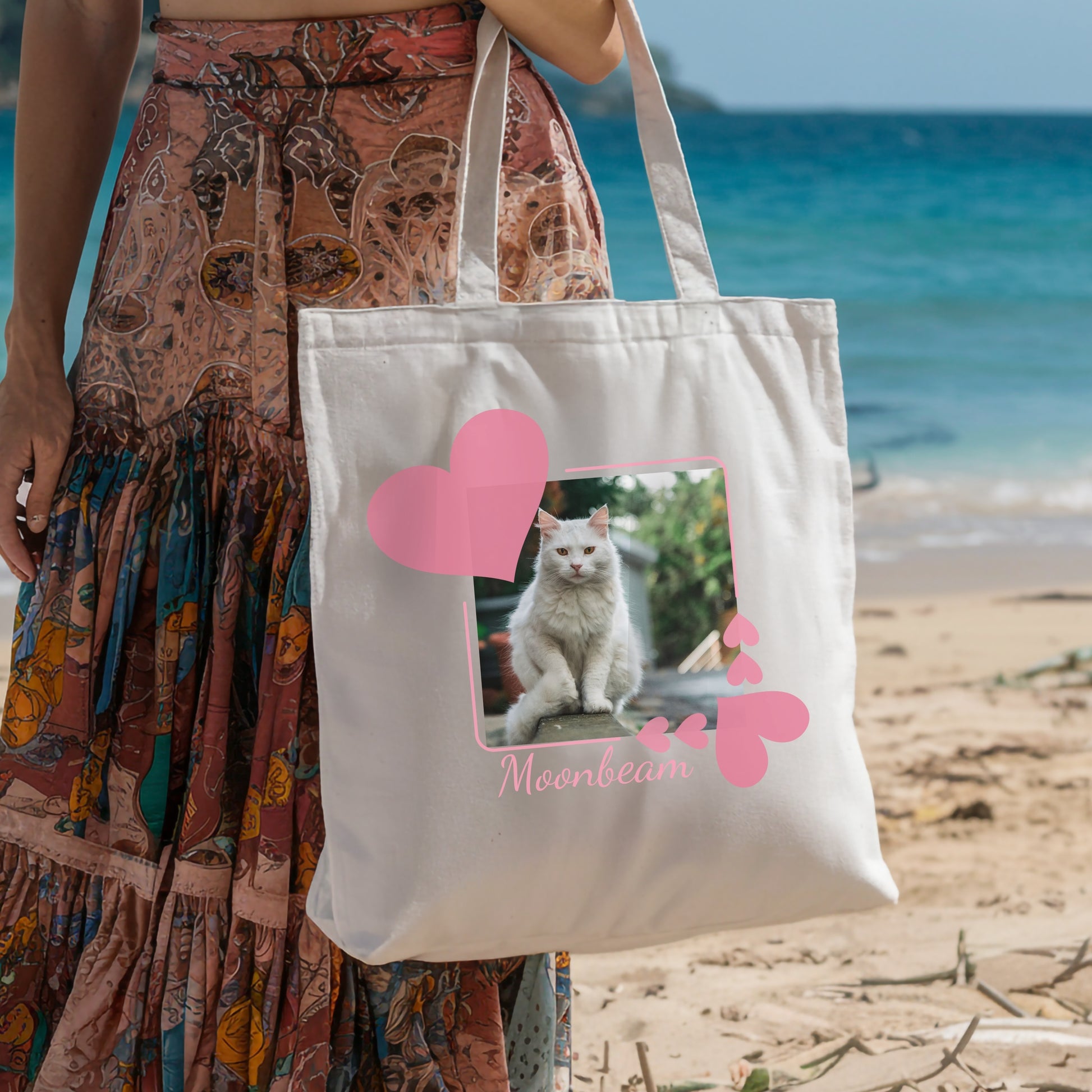 Person walking on the beach holding a tote bag with a picture of her cat in a pink  heart frame on it. 