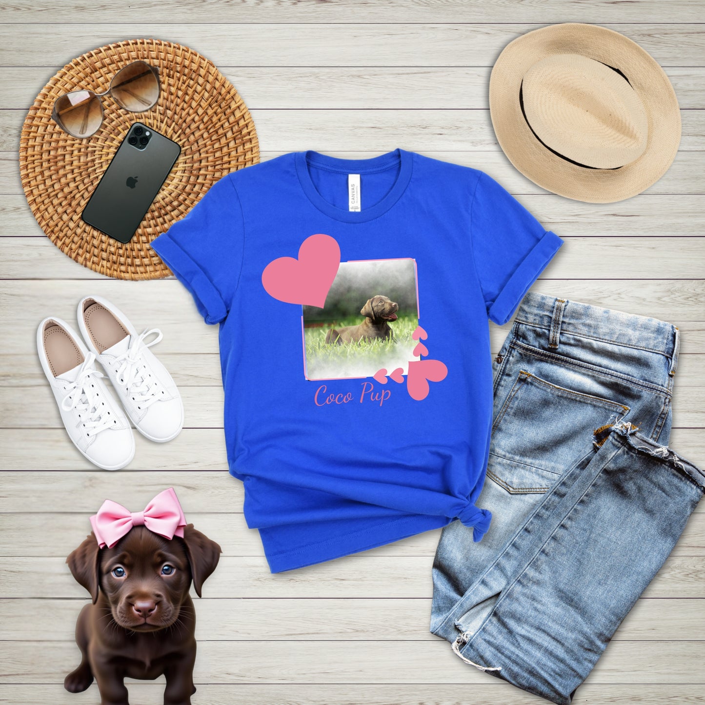 Blue t-shirt with a heart frame graphic that has a photo of a dog inside of it.  Below the frame is the dog's name, surrounded by a puppy, shoes, hat, and phone on a wooden surface.