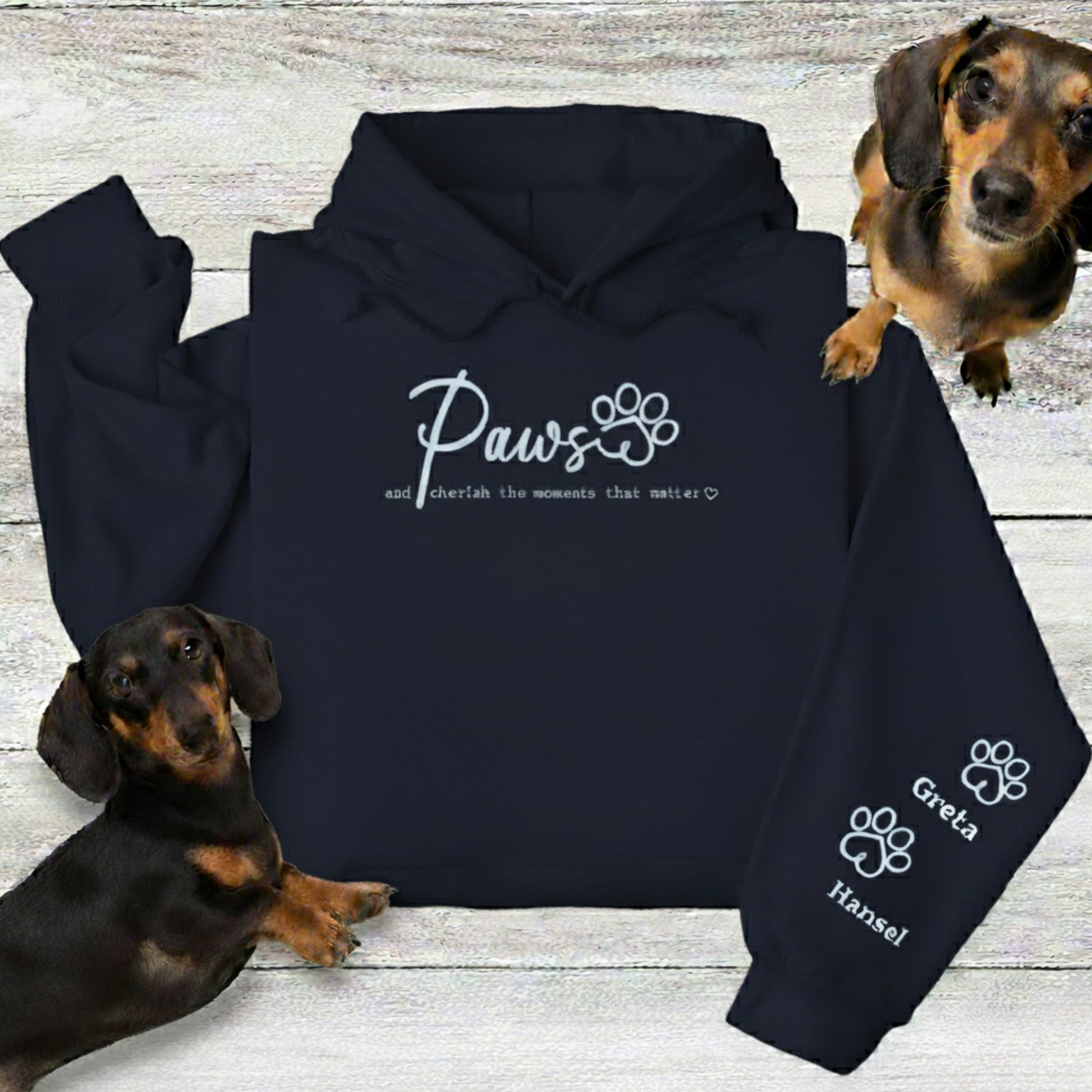 Personalise an embroidered Navu Blue hooded sweatshirt with up to four pets' names. The folded hoodie is shown with 2 Dachshunds on a wooden surface.