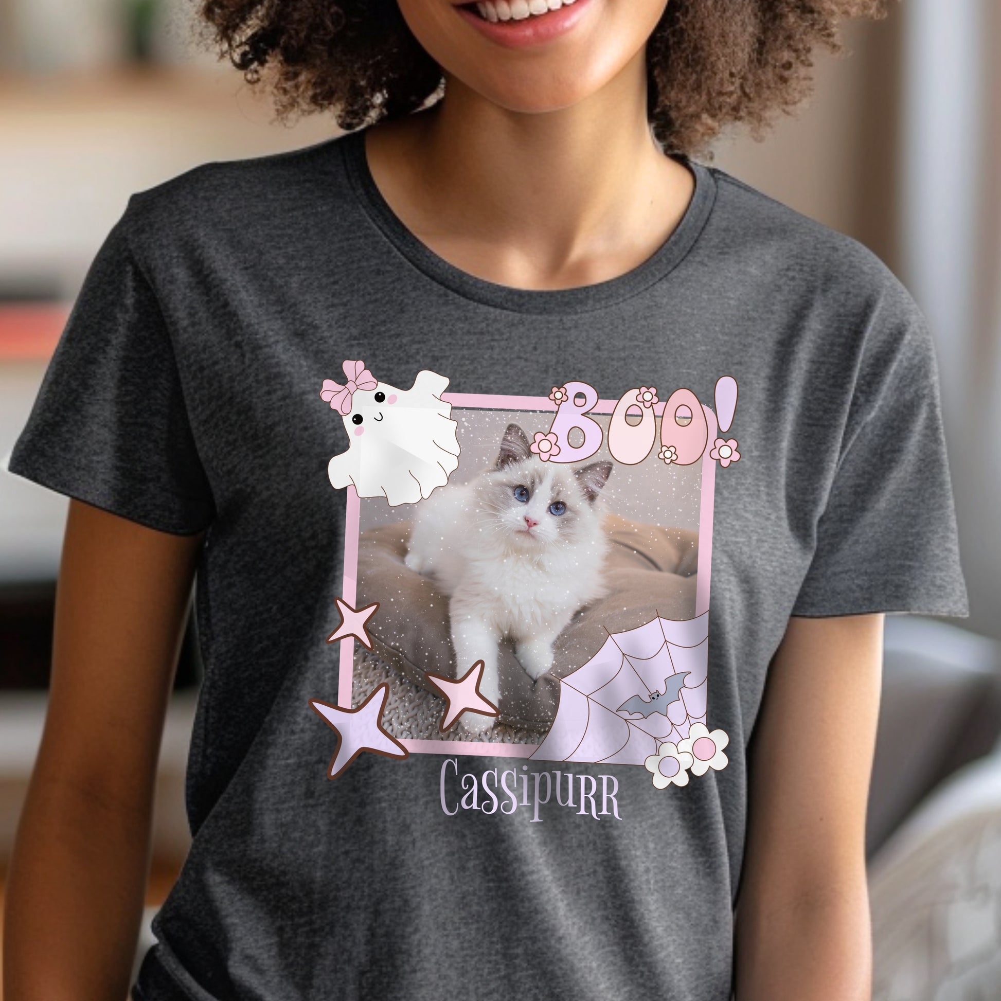 A woman wearing a dark heather gray t-shirt with a Coqueete Pink Halloween picture frame graphic that has a Ragdoll Cat inside of it. Below the frame in spooky font is the name, 'Cassipurr.'