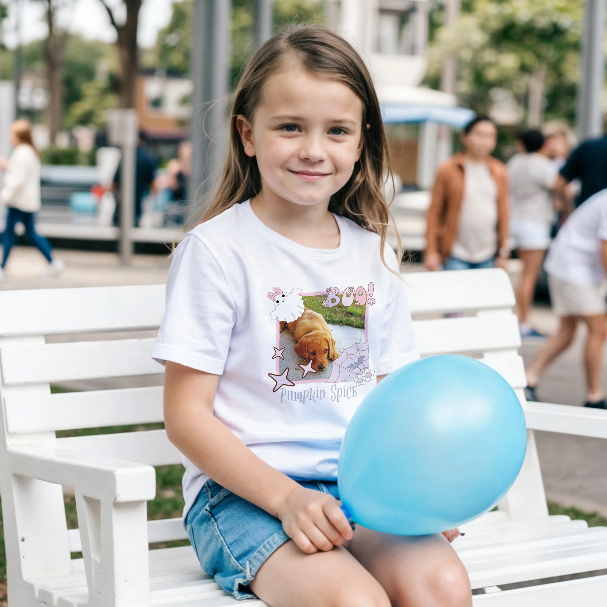 Young girl sitting on a bench holding a blue balloon, wearing a white t-shirt with a Coquette pink Halloween picture frame  graphic. The frame has a photo of a puppy and the text below, 'Pumpkin Spice.'