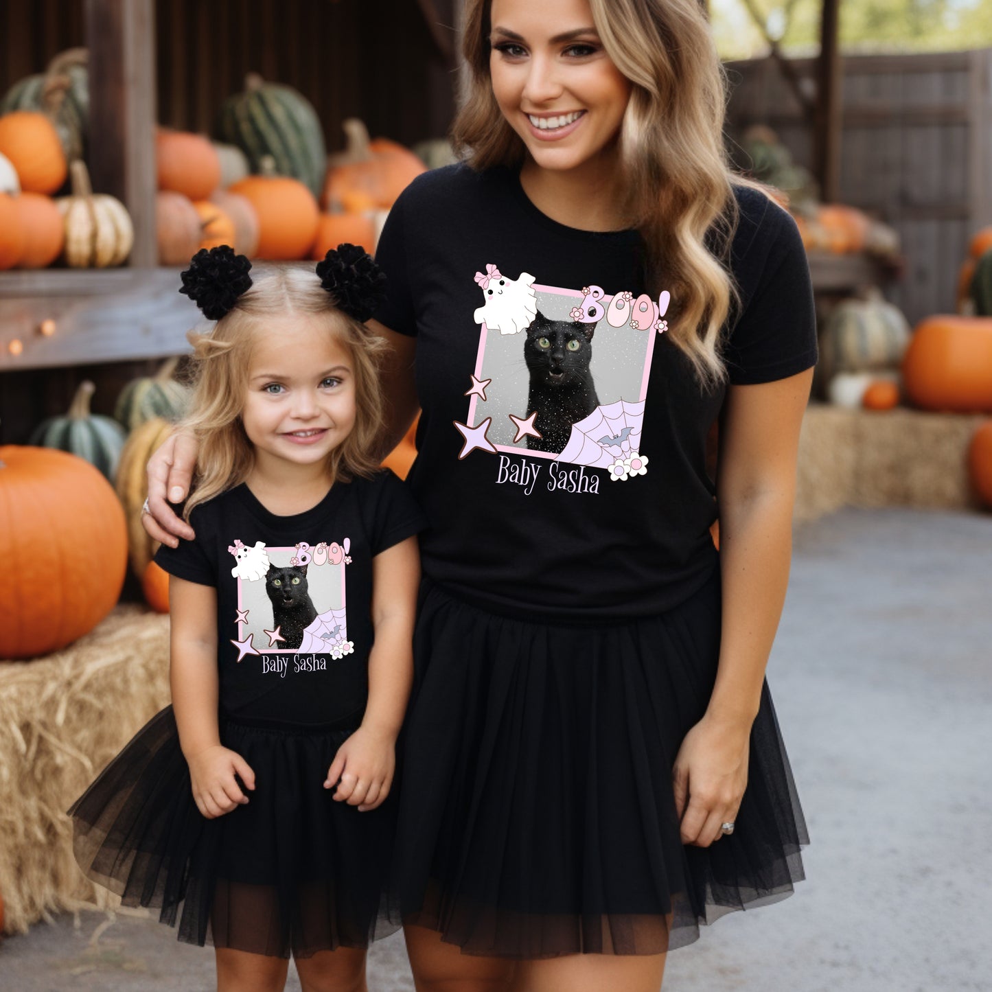 Woman and child wearing black t-shirts with a Coquette pink Halloween picture frame graphic with a black cat photo inside and text, Baby Sash' in a pumpkin patch.