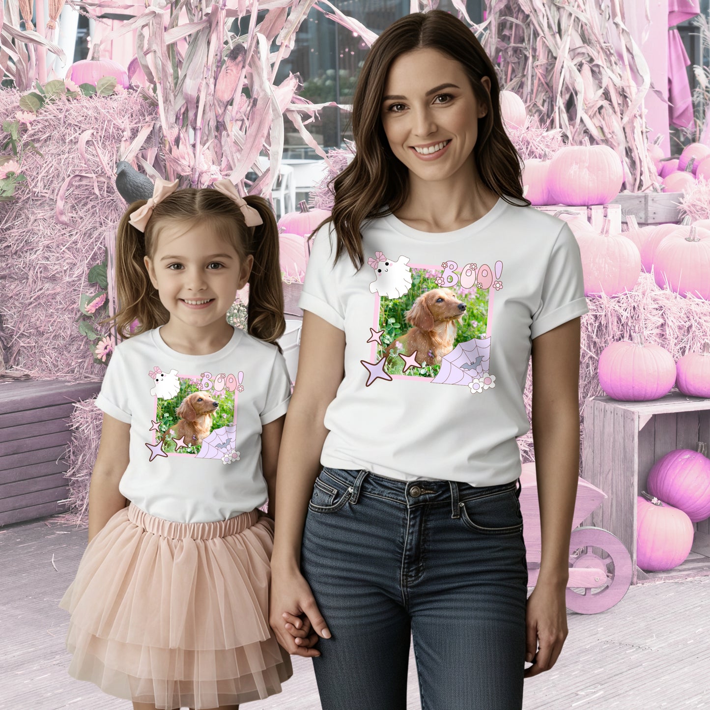 Mother and daughter wearing matching white t-shirts with a coquette pink Halloween picture frame  graphic containing a photo of a long haired Dashchund inside. They are standing in a whimsical pink pumpkin patch.