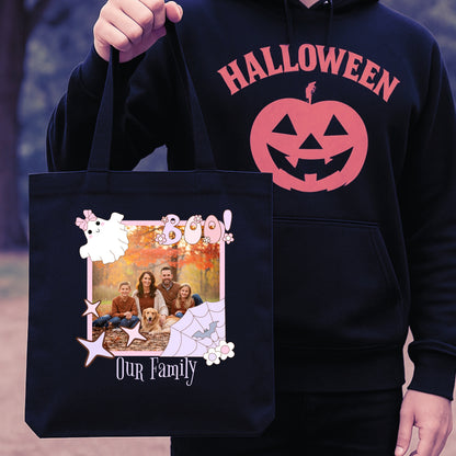 Person wearing a black hoodie with a Halloween design, holding a black tote bag with a family photo and 'Boo' text on a pink Halloween graphic.