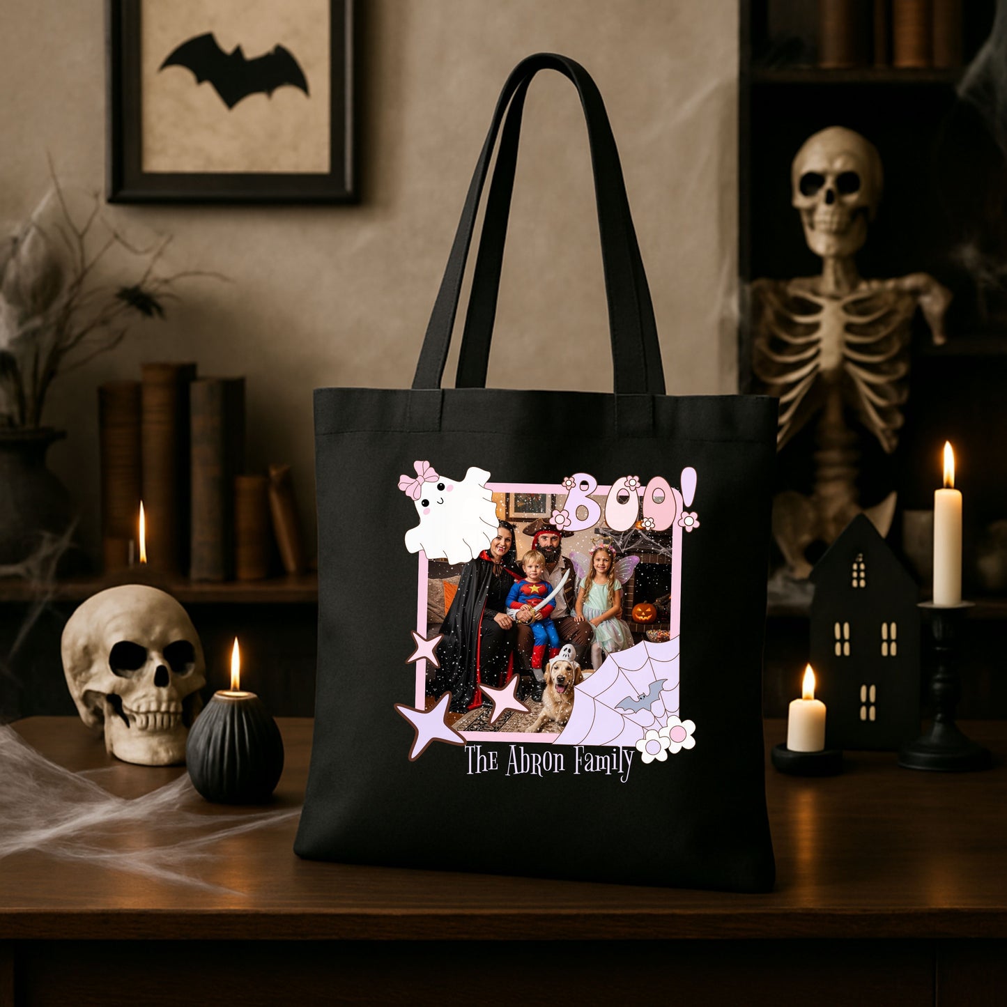 Black tote bag with a family photo design inside a Coquette pink Halloween picture frame graphic sitting on a Halloween-themed table with candles and a skeleton.