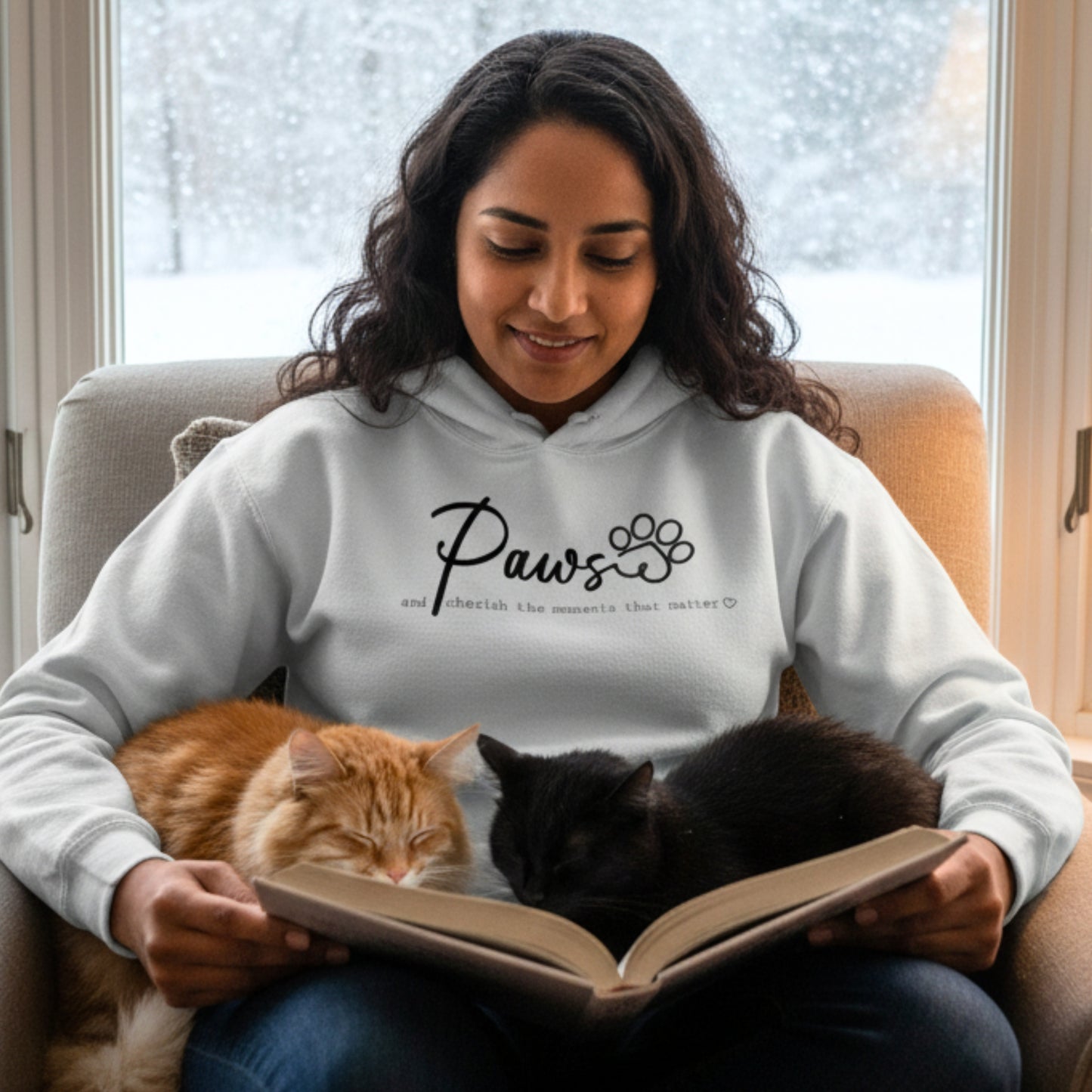Grey Hoodie shirt with custom embroidery. Embroidered text on the front of the Hooded Sweatshirt reads, 'Paws and cherish the moments that matter.' Life style photo of a woman in the embroidered sweatshirt enjoying a cozy moment inside with a book and her cats