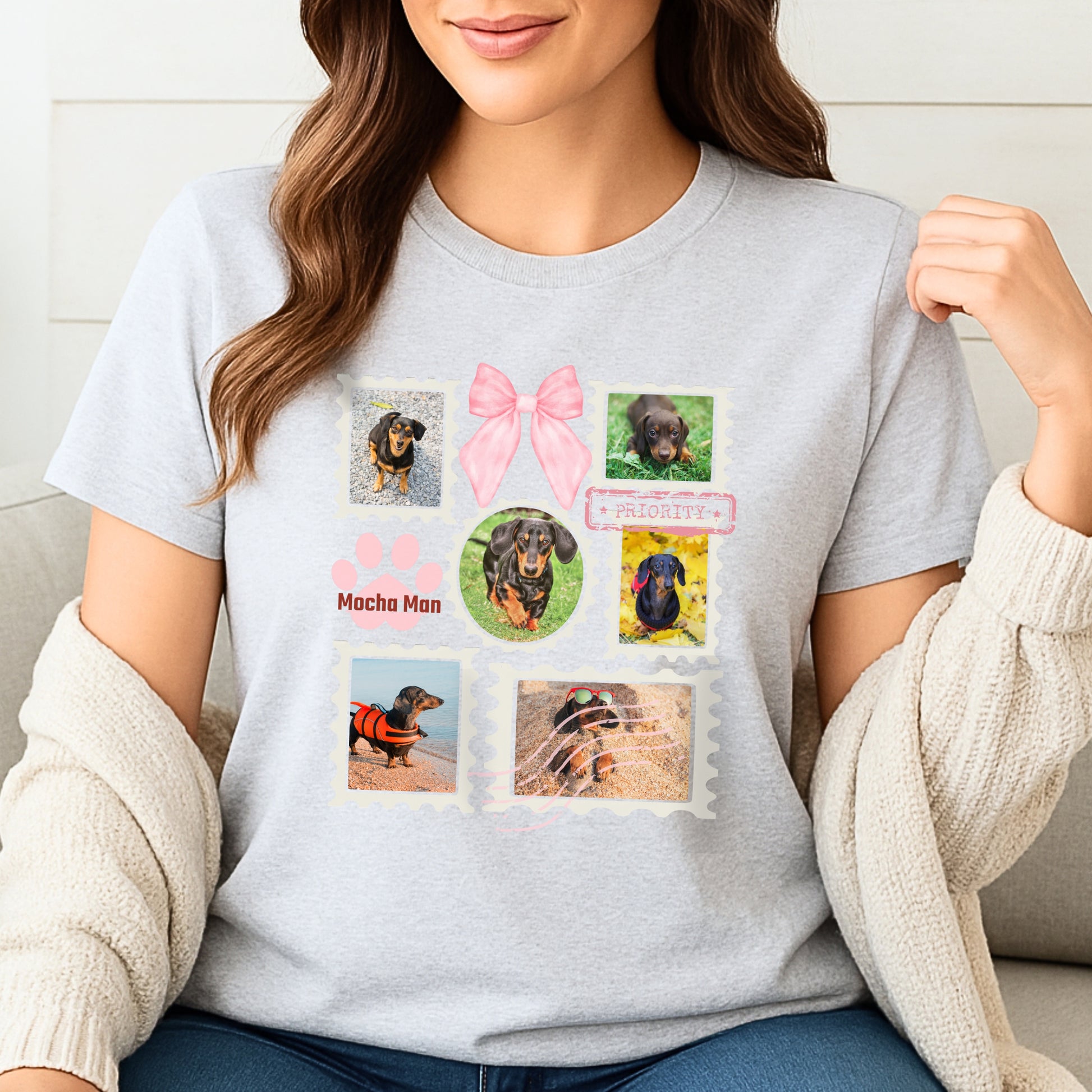 Woman wearing a light grey t-shirt with colorful graphic designs of her dogs as postage stamps and a pink coquette bow. She is sitting on a sofa.