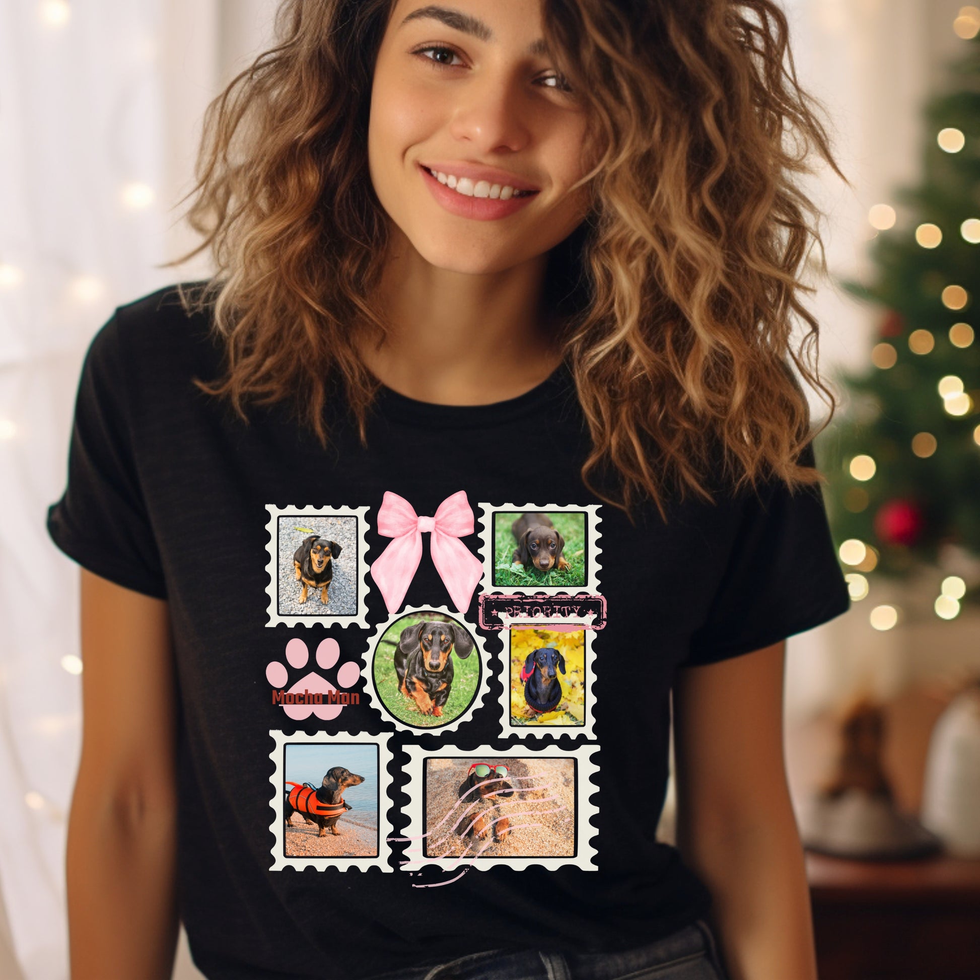 Woman wearing a black t-shirt with colorful graphic designs of her dogs as postage stamps and a pink coquette bow, Christmas tree in the background.