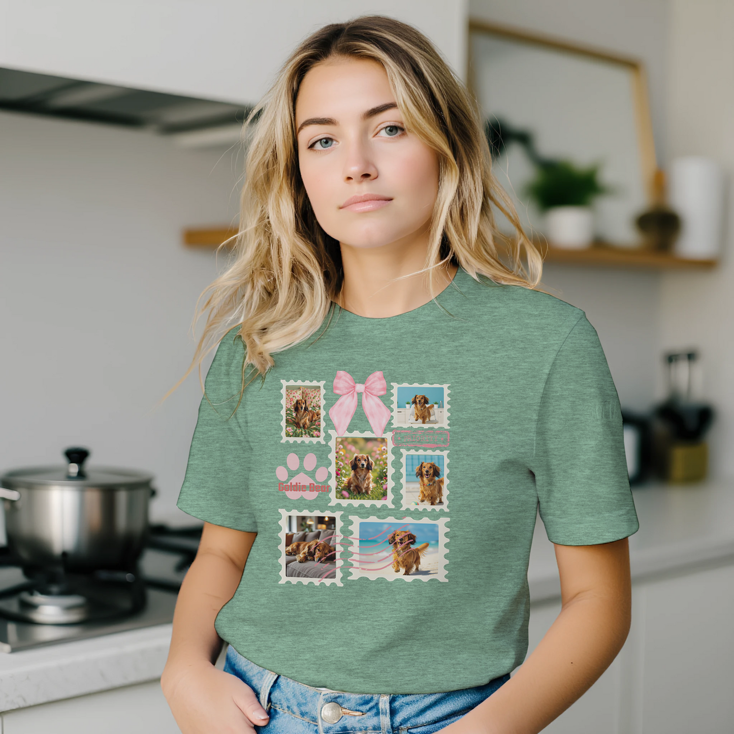 Woman wearing a heather mint green t-shirt with colorful graphic designs of her dogs as postage stamps and a pink coquette bow. She is standing in her kitchen.