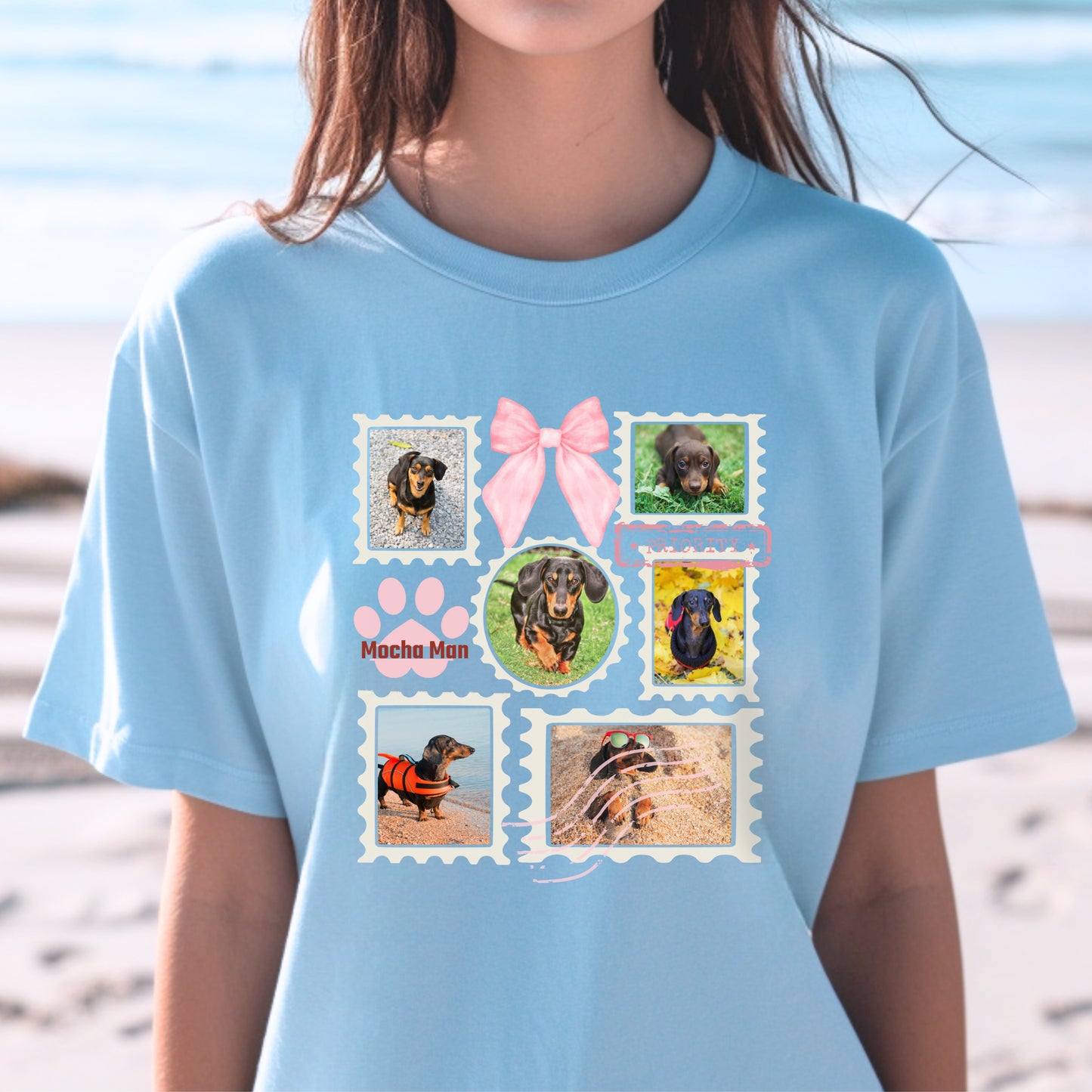Woman wearing a light blue t-shirt with colorful graphic designs of her dogs as postage stamps and a pink coquette bow. She is at the beach.