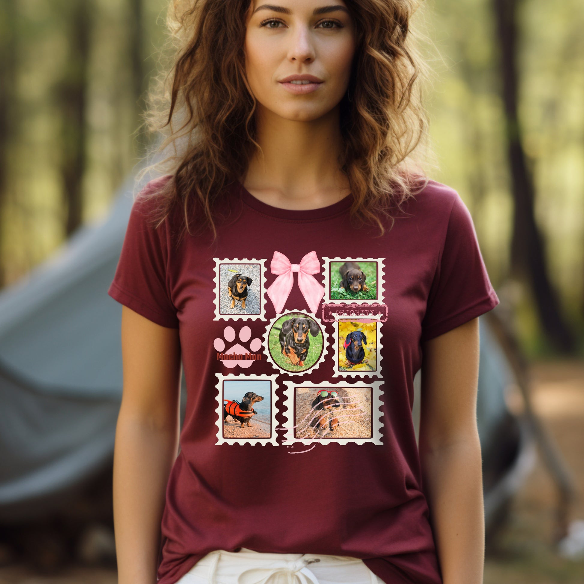 Woman wearing a maroon t-shirt with colorful graphic designs of her dogs as postage stamps and a pink coquette bow. She is camping in the woods.